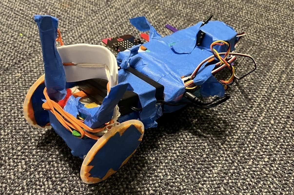 Prototype all-terrain robot with lid wheels, tape, and a micro:bit controller on a carpeted floor.